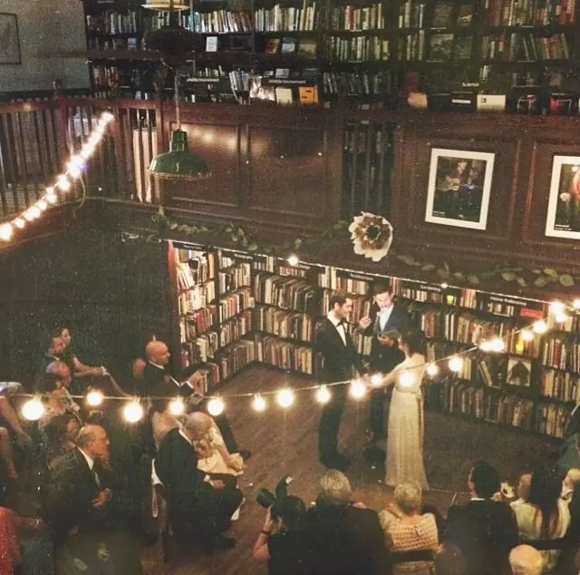 A couple exchanging vows before seated guests in a bookstore lined with tall shelves and string lights